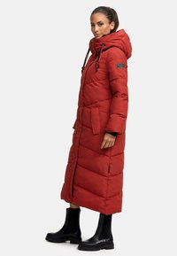 Manteau matelassé long et rouge avec capuche, doté de cordons élastiques, de poches latérales et d'un motif matelassé. Porté avec des bottines noires à la cheville.