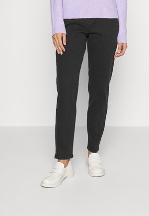 Osoba ubrana w czarne jeansy o prostych nogawkach, białe buty wsuwane i lawendowy sweter z długim rękawem stojąca na szarej podłodze.