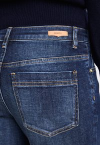 Jeans de mezclilla azul oscuro con una etiqueta de cuero en la cintura, costuras naranjas y un diseño clásico de cinco bolsillos.