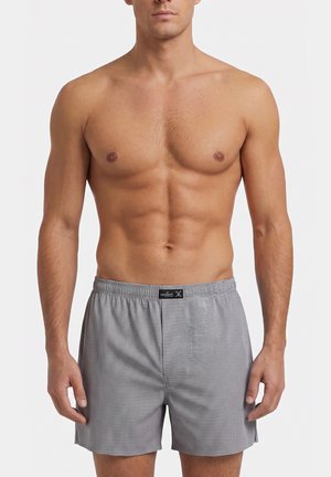 Oberkörperfreier muskulöser Mann trägt graue karierte Boxershorts und steht vor einem schlichten weißen Hintergrund mit entspannt an den Seiten hängenden Armen.