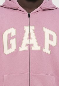 Ροζ φούτερ με φερμουάρ και λογότυπο "GAP" σε ανοιχτό μπεζ. Διαθέτει κουκούλα, μπροστινές τσέπες και μαλακή υφή. Το φερμουάρ εκτείνεται στο κέντρο.