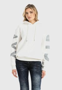 White hoodie with gray graphic accents on sleeves, drawstrings, and ribbed hem. Paired with dark blue distressed jeans.