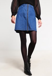 Jupe en jean avec une fermeture éclair frontale, présentant une forme classique en A, de couleur bleue et un style décontracté, portée avec des collants noirs et des bottines.