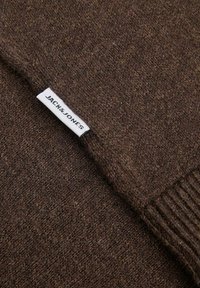 Tejido de punto marrón con una pequeña etiqueta blanca que dice "JACK & JONES" colocada cerca de un borde acanalado.