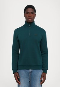 Tmavozelená mikina na zips s vysokým golierom, s čiernym logom na hrudi a rebrovanými manžetami, skombinovaná so svetlomodrými džínsami.