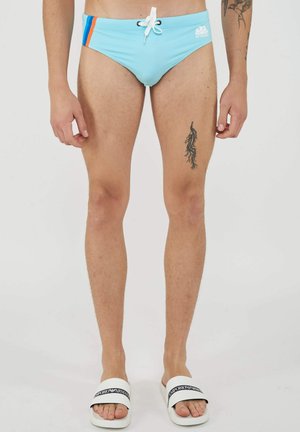 Man draagt lichtblauwe zwembroek met oranje en blauwe strepen aan de zijkant, wit koord en witte Emporio Armani slippers.