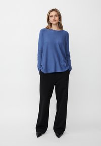 Frau in einem locker sitzenden blauen Langarmoberteil, schwarzen weit geschnittenen Hosen und schwarzen spitzen Schuhen, die mit Händen in den Taschen vor einem weißen Hintergrund steht.