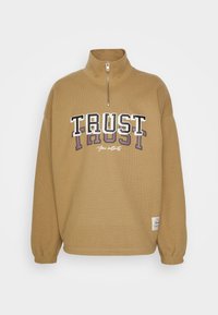 Brun lynlåstrøje med ribbet tekstur. Har et markant "TRUST"-motiv i sort og hvid, med accenter i lilla. Elastiske ærmekanter.