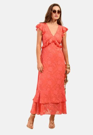 Kvinde iført koralfarvet blomster-middellang kjole med flæseærmer og kant, gyldne armbånd, holder en gylden håndtaske og har solbriller på.