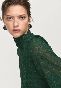 Blusa verde con estampado, de cuello alto y tejido drapeado. Pendientes circulares negros añaden contraste. La textura parece suave y ligera.