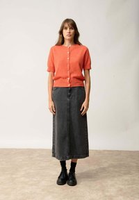 Korallfärgad kortärmad cardigan med knappar, i kombination med en lång svart denimkjol och svarta platåskor, med en neutral bakgrund.