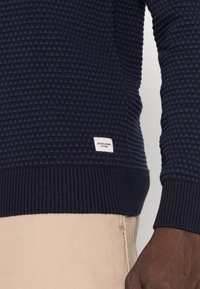 Marinblå texturerad stickad tröja med ribbade mudd och nederkant, med en liten etikett med "JACK & JONES" i vitt längst ner.