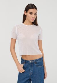 Top in rete bianco, corto, decorato con strass sparsi, con maniche corte e scollo rotondo, abbinato a jeans a vita alta.