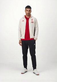 Casaco branco com fecho éclair, com logótipo do Liverpool em vermelho, camisola vermelha e calças de treino pretas. Ténis desportivos brancos com detalhes em vermelho. Texturas suaves.