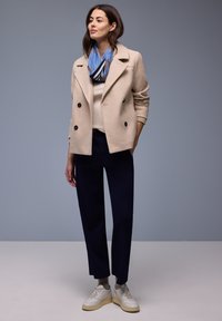 Mujer de pie con abrigo beige cruzado, bufanda azul, pantalones azul marino, calcetines grises y zapatillas blancas frente a un fondo gris.