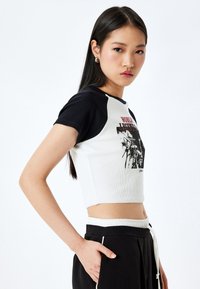 Geribbelde crop top in wit en zwart, met korte zwarte mouwen en een grafisch ontwerp aan de voorkant. Gecombineerd met zwarte broeken.