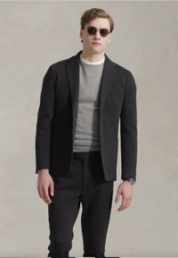 Blazer noir ajusté sur un pull gris, associé à un pantalon noir. Les accessoires incluent des lunettes de soleil foncées et une montre-bracelet. Textures lisses.