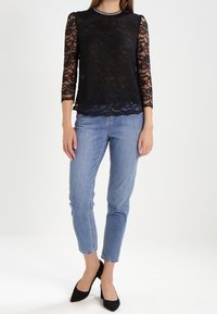 Top in pizzo nero con maniche a tre quarti, scollatura rotonda e orlo a scallop, abbinato a jeans azzurri chiari e tacchi neri a punta.