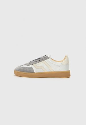 Low-top sneaker met een witte en beige leren bovenkant, grijze suède neus en veters en een natuurlijke rubberen zool, zijaanzicht.