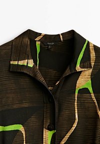 Chemise à col avec des motifs abstraits noirs, marron, beige et vert vif et des boutons noirs sur le devant.