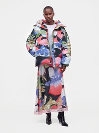 Femme portant une doudoune à motif abstrait coloré et une longue jupe assortie avec des bottines à talons noires, debout sur un fond uni.