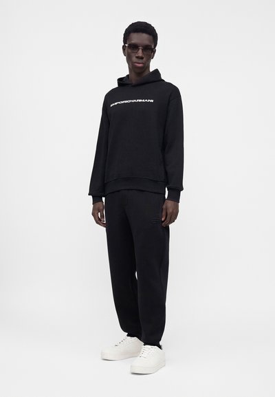 Мъж, облечен с черен суитшърт Emporio Armani, черен панталон, бели маратонки и тъмни слънчеви очила, стои срещу светъл фон.