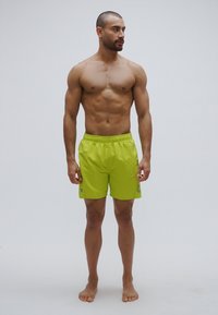 Pantalones cortos deportivos verde lima con cintura elástica y detalle de logotipo, fabricados con tejido ligero, modelados por un hombre de complexión musculosa.