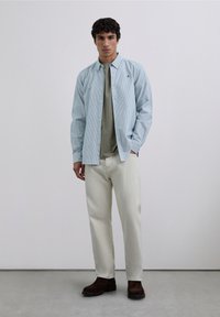 Lichtblauw gestreept overhemd over een groen T-shirt, gecombineerd met roomwitte broek en bruine schoenen. Eenvoudig ontwerp en zachte stoftexturen.