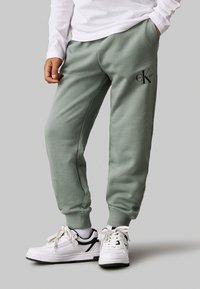 Pantaloni della tuta verdi realizzati in tessuto morbido, con vita elastica e caviglie a polsino. Presentano un logo nero sulla coscia sinistra.