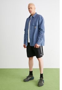 Blå långärmad skjorta med framfickor, kombinerad med svarta shorts med vita ränder. Bärs med gråa sportskor och svarta strumpor.