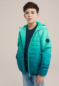 Vadderad jacka i en ombre teal-färg, med dragkedjestängning, huva och ett texturerat tyg med en rund patch på vänster arm.
