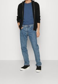 Svart ribbad zip-up tröja, blå t-shirt, ljusblå denimjeans och svarta sneakers med vit sula, stående mot en neutral bakgrund.