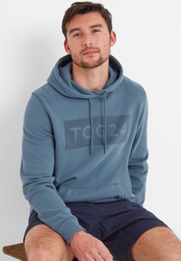 Blue hoodie with a kangaroo pocket and drawstring hood. Features a large printed logo "TOG24" in a darker shade on the front.