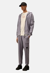 Helle graue Kapuzenpullover mit seitlichem Reißverschlussfach, kombiniert mit passenden Slim-Fit-Hosen, die über eine seitliche Cargo-Tasche und schwarze Akzente verfügen.
