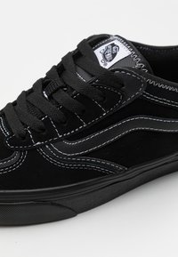 Svart semsket sneakers med hvite sømmer, teksturerte detaljer og perforerte elementer på sidene. Har en flat svart gummisåle.