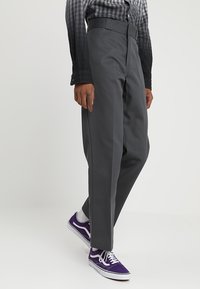 Person som bär grå raka byxor, en svart och grå rutmönstrad skjorta, vita strumpor och lila sneakers som står framför en enfärgad bakgrund.