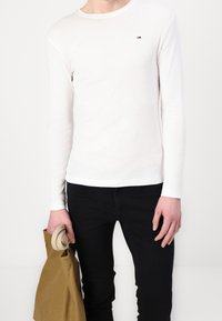 Persona vistiendo una camisa blanca de manga larga con un pequeño logo en el pecho, pantalones negros y sosteniendo un objeto enrollado beige y una bolsa marrón.