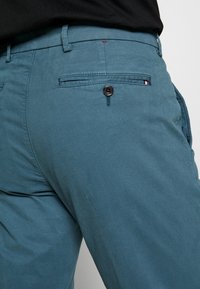 Teal katoenen broek met een slank ontwerp, voorzien van een achterzak met een zwarte knoop en een rode accentnaad. Glad textuur is zichtbaar.