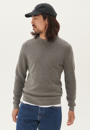 Maglione grigio a maglia con collo rotondo e polsini a costine, indossato sopra una camicia bianca, abbinato a jeans blu e un berretto nero.