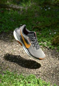 Gray athletic shoe with a breathable mesh upper, orange accents, and textured sole, suspended above a gravel and grass surface.