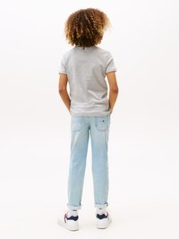 Maglietta grigia a maniche corte, jeans di denim azzurri, sneakers bianche con accenti blu e rossi, in piedi con le mani dietro la schiena, riccioli testurizzati.