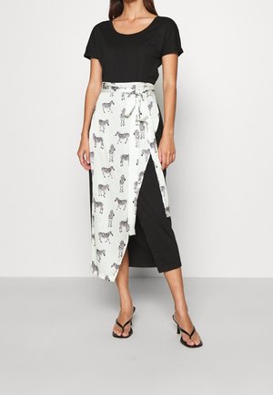 Frau trägt ein schwarzes Kleid mit kurzen Ärmeln und einem weißen Wickelrock mit schwarzen Zebrastreifen, kombiniert mit schwarzen Sandalen mit Absatz.