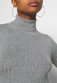 Gros plan d'une personne portant de grandes boucles d'oreilles en or et un pull à col roulé gris côtelé.