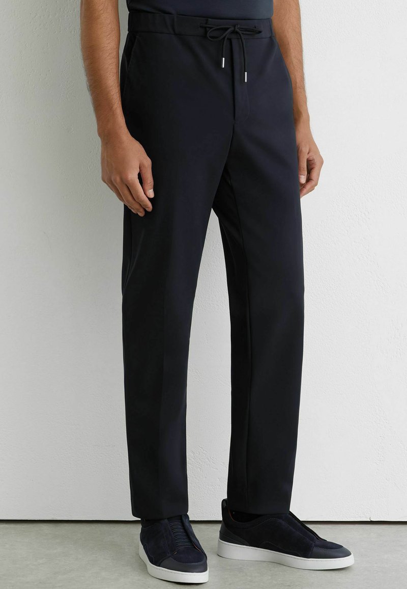 Hombre que lleva pantalones negros con cordón y zapatillas negras con suelas blancas, de pie contra una pared blanca con textura.