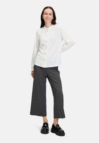Chemise blanche à boutons avec un motif géométrique, associée à un pantalon gris taille haute et à jambes larges, et à des chaussures à plateforme noires.