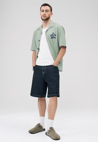 Light green short-sleeve shirt with blue text, white tank top, dark denim shorts, and beige slip-on shoes paired with white socks.