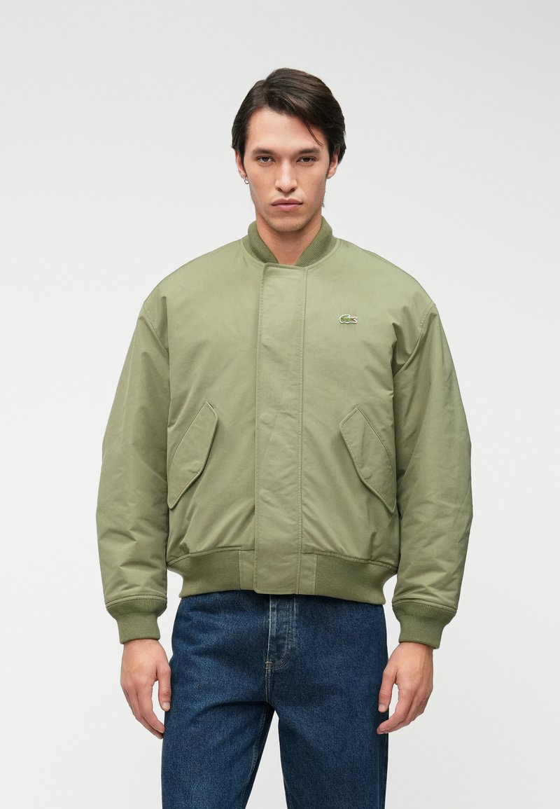 Πράσινο μπομπέρ μπουφάν με ψηλό γιακά, μπροστινές τσέπες, ριμπ μανσέτες και στρίφωμα, φτιαγμένο από ομαλό, ελαφρύ ύφασμα. Λογότυπο Lacoste στο στήθος.