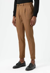 Bruine wollen broek met een getailleerde pasvorm, voorzien van een voorplooi en omgeslagen pijpen, gecombineerd met zwarte leren loafers.