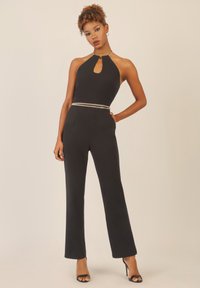 Elegant jumpsuit nero con scollatura a chiave, accentuato da una cintura in catena argentata, design aderente e pantaloni a gamba larga. Indossato con tacchi neri.