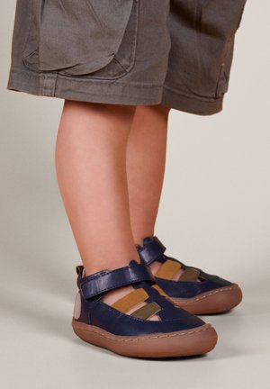 Zapatos de cuero azul marino con diseño de correas cruzadas; cuentan con acentos en marrón y oliva y suela flexible de goma marrón para mayor comodidad.